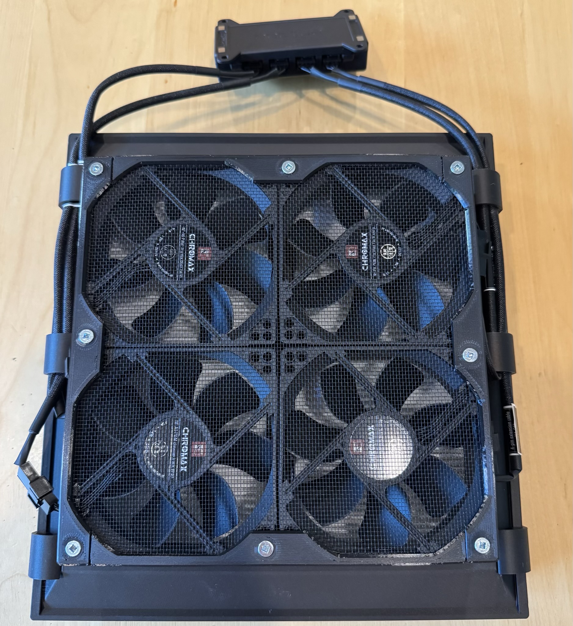 Four fans are behind a screen, attached to a black 3-d printed cowl with filters on the back side, just visible between the fan blades. Four wires snake up the sides and meet at a black box at the top.